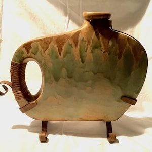 May include: A ceramic flask with a distinctive design, featuring a large handle and a flat, rounded body. The flask is supported by a metal stand. The colours include shades of brown, green, and tan, creating a rustic aesthetic.