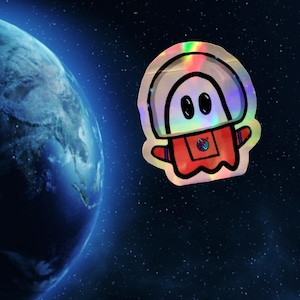 May include: A holographic sticker featuring a cartoon astronaut in a red spacesuit with a white helmet. The sticker has a rainbow sheen and is set against a backdrop of a blue planet and a starry night sky.