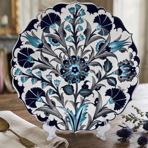May include: A round decorative plate with a scalloped edge featuring a symmetrical floral pattern in shades of navy blue, light blue, and grey on a white background, displayed on a clear plastic stand with a central circular flower motif.