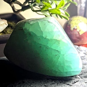 May include: A polished, rounded, green gemstone with a textured surface. The stone is a vibrant shade of green, with lighter areas that catch the light. The gemstone is displayed on a dark, textured surface, with a small plant in the background.