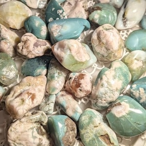 May include: A selection of polished gemstones in teal, cream, and brown hues. The stones are of varying sizes and shapes, with smooth, rounded surfaces. Some have a marbled appearance, while others are more solid in colour. The gemstones are arranged on a white, lacy surface.