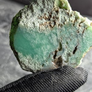 May include: A close-up of a rough-cut gemstone with a vibrant, sea-green hue. The stone has a textured, uneven surface with areas of lighter green and brown. The gemstone is held by a black fabric.