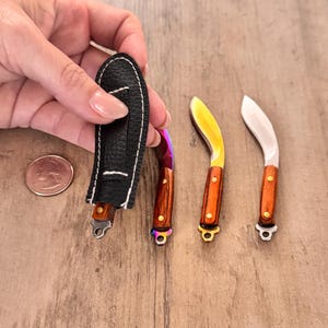 May include: Miniature knives with wooden handles and curved blades. One knife is in a black leather sheath. The blades have a rainbow, gold, and silver finish. A US quarter is next to the knives.