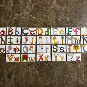 Pode incluir: Um conjunto de cartões retangulares apresentando o alfabeto inglês de A a Z. Cada cartão exibe uma letra maiúscula e minúscula ilustrada criativamente como animais, objetos ou personagens em várias cores, dispostos em quatro linhas.