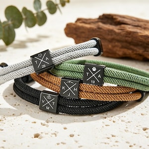 Op de afbeelding: Een verzameling touwarmbanden in grijs, groen, bruin en zwart. Elke armband heeft een zwarte vierkante bedel met initialen en symbolen. De armbanden liggen op een lichtgekleurd oppervlak.