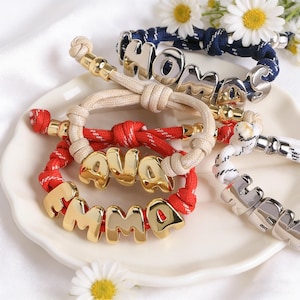 Puede incluir: Pulseras personalizadas con letras doradas y plateadas que deletrean nombres y la palabra "HOME". Las pulseras tienen cordones rojos, azul marino, blancos y beige. Las pulseras se exhiben en un plato blanco con margaritas.