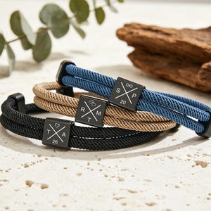 Peut inclure: Trois bracelets en corde noire, beige et bleu, chacun avec un fermoir carré noir. Les fermoirs présentent du texte et des symboles blancs. Les bracelets sont présentés sur une surface texturée blanc cassé.