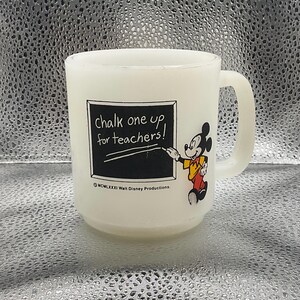 K&ouml;nnte beinhalten: Wei&szlig;er Glasbecher mit Henkel, mit Mickey Mouse und dem Schriftzug "chalk one up for teachers!". Mickey ist in seiner ikonischen Kleidung abgebildet und zeigt auf eine Tafel. Der Becher ist wahrscheinlich ein Vintage-Artikel.
