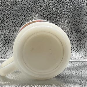K&ouml;nnte beinhalten: Eine wei&szlig;e Keramiktasse mit Henkel, von unten betrachtet. Der Rand hat ein dekoratives Band mit orangefarbenen, roten und gelben Akzenten. Die Tasse steht vor einem strukturierten, silbernen Hintergrund.