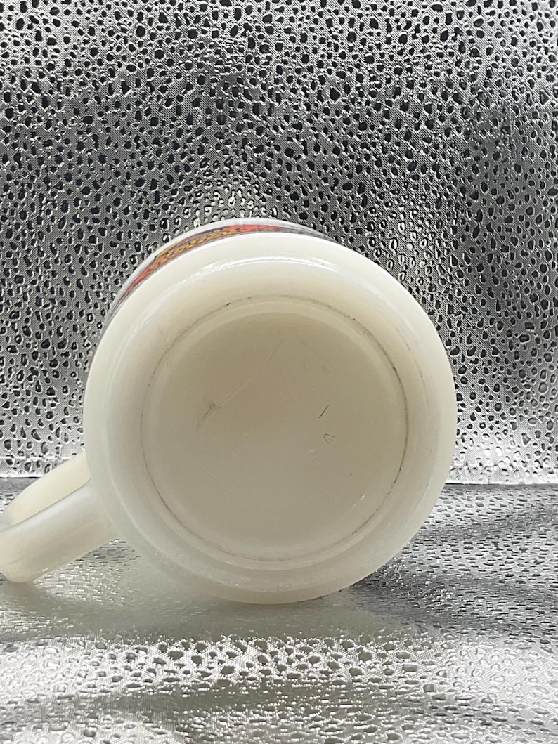 K&ouml;nnte beinhalten: Eine wei&szlig;e Keramiktasse mit Henkel, von unten betrachtet. Der Rand hat ein dekoratives Band mit orangefarbenen, roten und gelben Akzenten. Die Tasse steht vor einem strukturierten, silbernen Hintergrund.