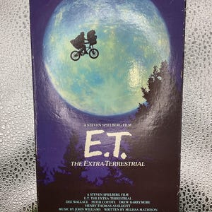 May include: A VHS tape for the movie "E.T. The Extra-Terrestrial." The cover features a silhouette of a figure on a bicycle in front of a large moon. The title and credits are displayed in white and yellow text against a dark blue background.