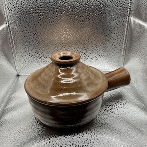 May include: A brown ceramic casserole dish with a lid and a handle. The dish is a warm brown colour with darker brown accents. The lid has a small opening at the top. The handle is a cylindrical shape.
