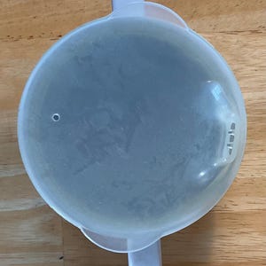 Puede incluir: Una tapa de pl&aacute;stico transparente con dos asas y un peque&ntilde;o agujero. La tapa es redonda y tiene una forma ligeramente abovedada. Las asas son rectangulares y se extienden desde los lados de la tapa. La tapa est&aacute; sobre una superficie de madera.