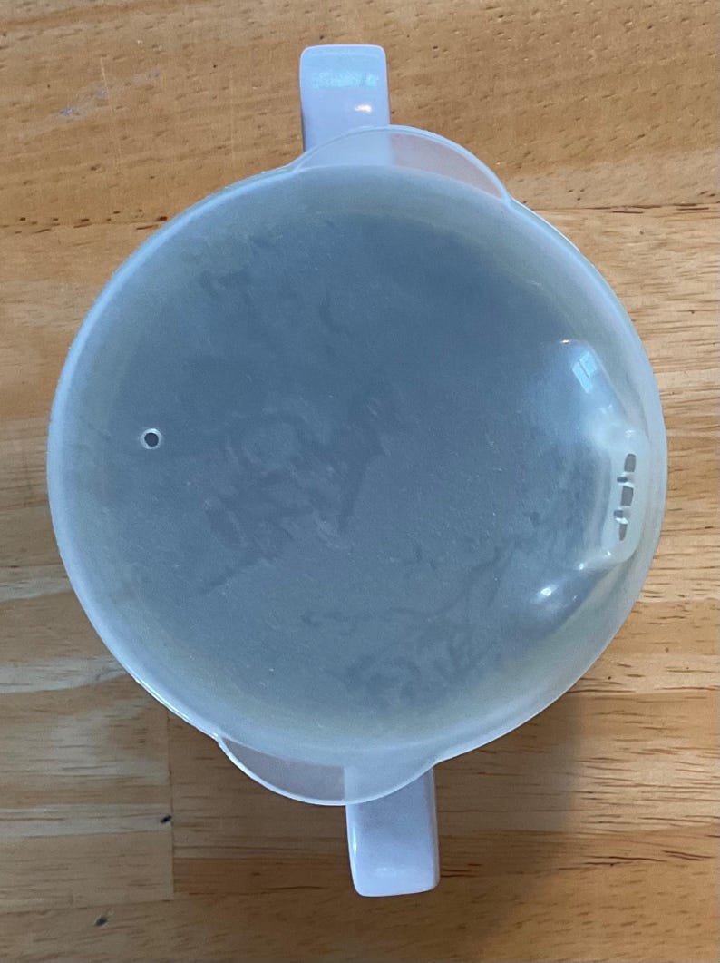 Puede incluir: Una tapa de pl&aacute;stico transparente con dos asas y un peque&ntilde;o agujero. La tapa es redonda y tiene una forma ligeramente abovedada. Las asas son rectangulares y se extienden desde los lados de la tapa. La tapa est&aacute; sobre una superficie de madera.