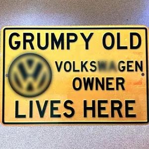 May include: A rectangular yellow metal sign with black text that reads "GRUMPY OLD VOLKSWAGEN OWNER LIVES HERE." The sign features the Volkswagen logo and a black border.