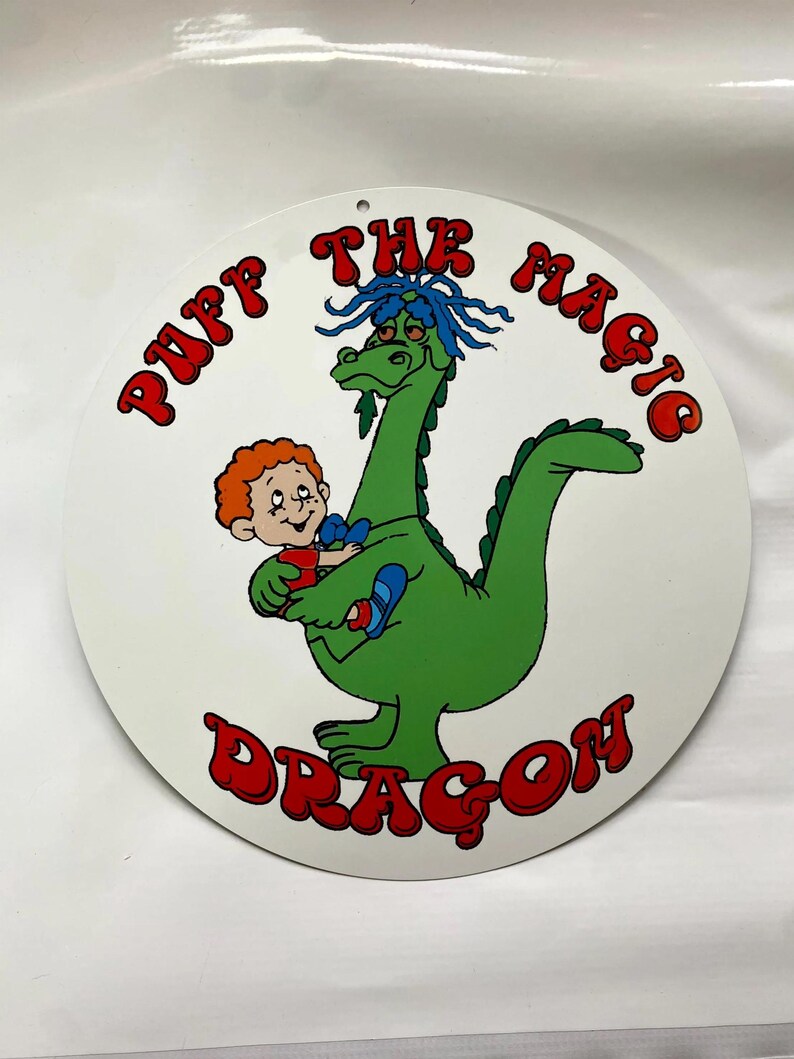Pu&ograve; includere: Cartello rotondo bianco con un'illustrazione a cartoni animati di un drago verde con i capelli blu che tiene in braccio un bambino. Il cartello riporta le parole "Puff The Magic Dragon" in lettere rosse sul bordo.