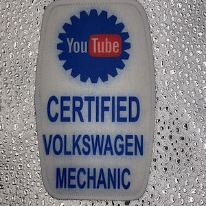 May include: A white patch featuring a blue cog design with the YouTube logo at its centre. Below the logo, the text reads "CERTIFIED VOLKSWAGEN MECHANIC" in blue. The patch is edged with a white border.