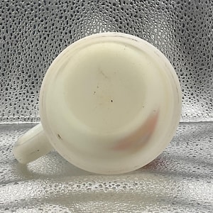 K&ouml;nnte beinhalten: Eine wei&szlig;e Keramiktasse mit Henkel, von unten betrachtet. Die Tasse hat eine abgerundete Form und einen kleinen Henkel an der Seite. Das Innere der Tasse ist sichtbar, und es gibt einige kleine Unvollkommenheiten auf der Oberfl&auml;che.