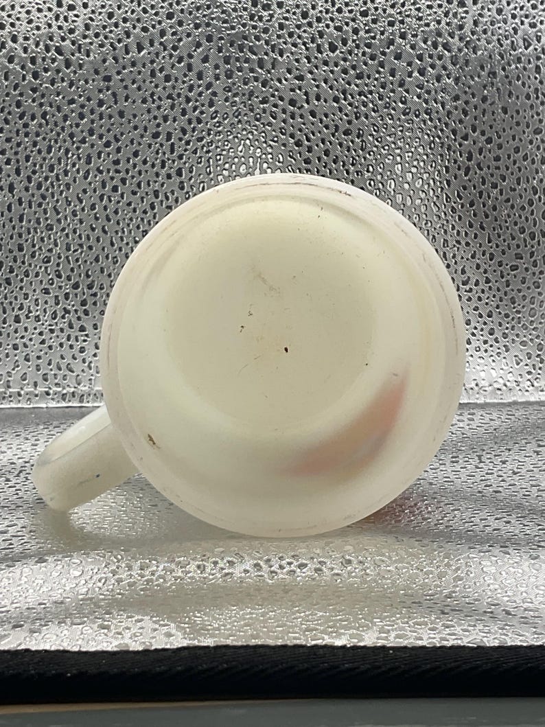 K&ouml;nnte beinhalten: Eine wei&szlig;e Keramiktasse mit Henkel, von unten betrachtet. Die Tasse hat eine abgerundete Form und einen kleinen Henkel an der Seite. Das Innere der Tasse ist sichtbar, und es gibt einige kleine Unvollkommenheiten auf der Oberfl&auml;che.