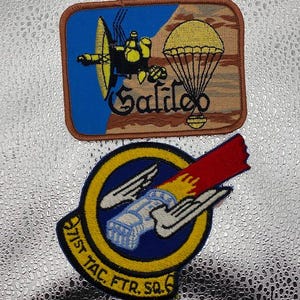 May include: Two embroidered patches. The top patch features a yellow spacecraft and the word "Galileo" on a blue and brown background. The bottom patch has a blue and yellow circular design with the text "971st TAC. FTR. SQ."