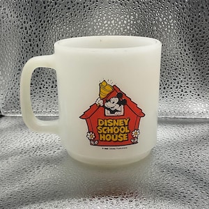 K&ouml;nnte beinhalten: Eine wei&szlig;e Keramik-Tasse mit Henkel. Die Tasse zeigt eine farbenfrohe Grafik von Micky Maus in einem roten Haus mit dem Text "DISNEY SCHOOL HOUSE". Eine Vintage-Sammeltasse.