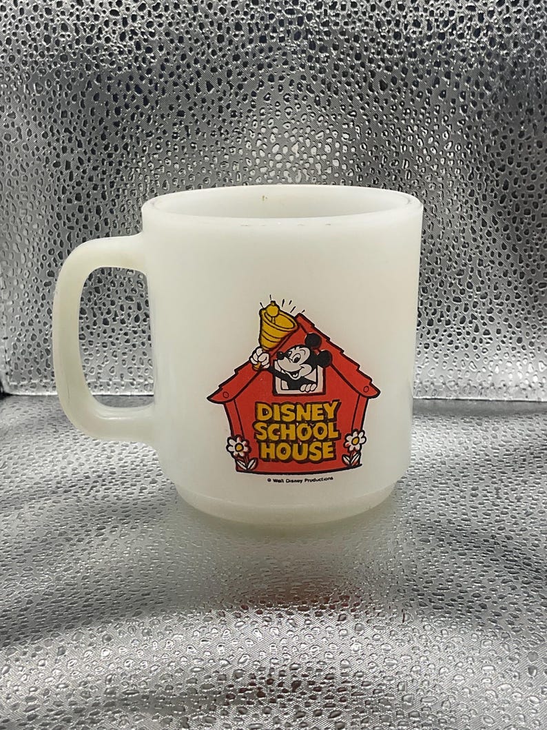 K&ouml;nnte beinhalten: Eine wei&szlig;e Keramik-Tasse mit Henkel. Die Tasse zeigt eine farbenfrohe Grafik von Micky Maus in einem roten Haus mit dem Text "DISNEY SCHOOL HOUSE". Eine Vintage-Sammeltasse.