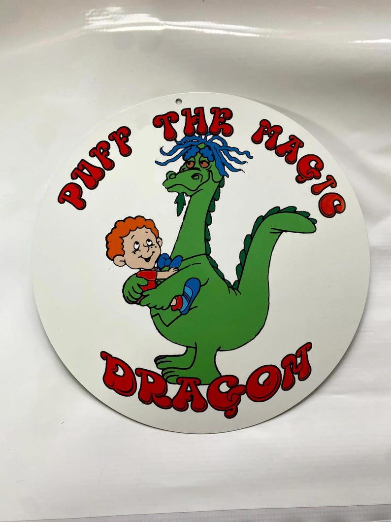 Pu&ograve; includere: Un cartello rotondo bianco con un'illustrazione a cartoni animati di un drago verde e un bambino. Il testo "Puff The Magic Dragon" &egrave; scritto in rosso attorno al bordo del cartello.