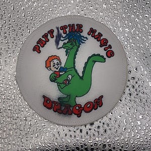 May include: A white circular patch featuring a green dragon with blue hair and a small boy. The text "PUFF THE MAGIC DRAGON" is arched around the top and bottom of the patch in red letters.