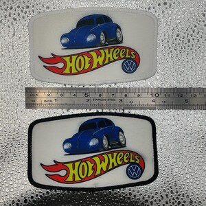 May include: Two rectangular patches featuring a blue cartoon car with the text "HOT WHEELS" in yellow and red. The patches have a white background and a black border. The Hot Wheels logo is also present.