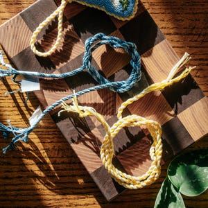 May include: Two braided cord bracelets, one in a variegated blue and white pattern and the other in a solid pale yellow, each featuring loose, frayed ends and a circular loop design, displayed as individual accessories for wrist or ankle wear.