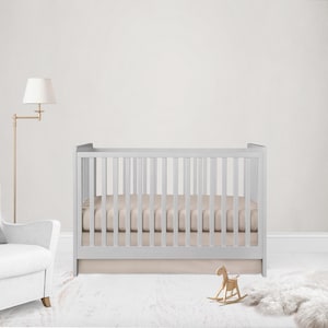 May include: A light gray crib with vertical slats, a beige mattress, and a matching skirt. A wooden rocking horse and a white rug are in front of the crib. A gold floor lamp and a white armchair are to the left.