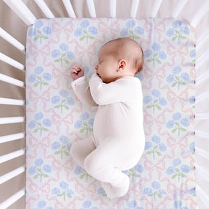 May include: A white crib sheet featuring a repeating pattern of light blue hydrangea flowers and soft pink ribbons. The sheet is fitted onto a white slatted crib mattress, providing a clean and simple surface for a sleeping infant in a white onesie.