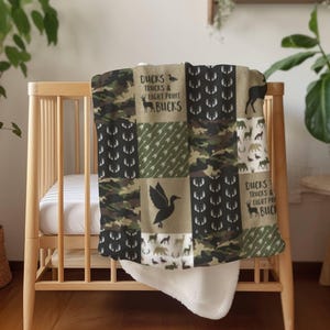 Puede incluir: Una manta de bebé de retazos con un patrón de camuflaje, siluetas de ciervos y el texto "Ducks, Trucks & Eight Point Bucks". La manta está sobre una cuna de madera, con un reverso blanco y suave.