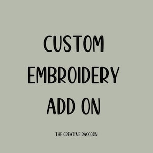 May include: A digital graphic on a muted green background. The text reads "CUSTOM EMBROIDERY ADD ON" in a bold, black, handwritten-style font. Below, in a smaller font, is "THE CREATIVE RACCOON".