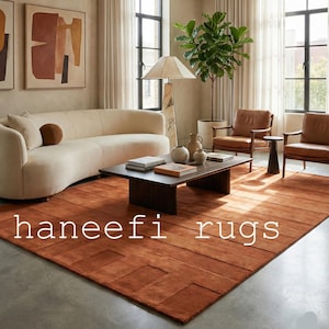 May include: A living room scene featuring a large, rust-colored rug with a geometric pattern. The rug is the focal point, complemented by a cream-colored sofa, brown leather chairs, and a dark wood coffee table. The text "haneefi rugs" is displayed on the rug.