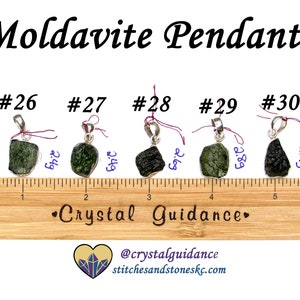 May include: Five silver pendants with green and black moldavite stones. The pendants are numbered 26 through 30 and are displayed on a wooden ruler. The stones are approximately 2.69 to 2.99 inches in size.