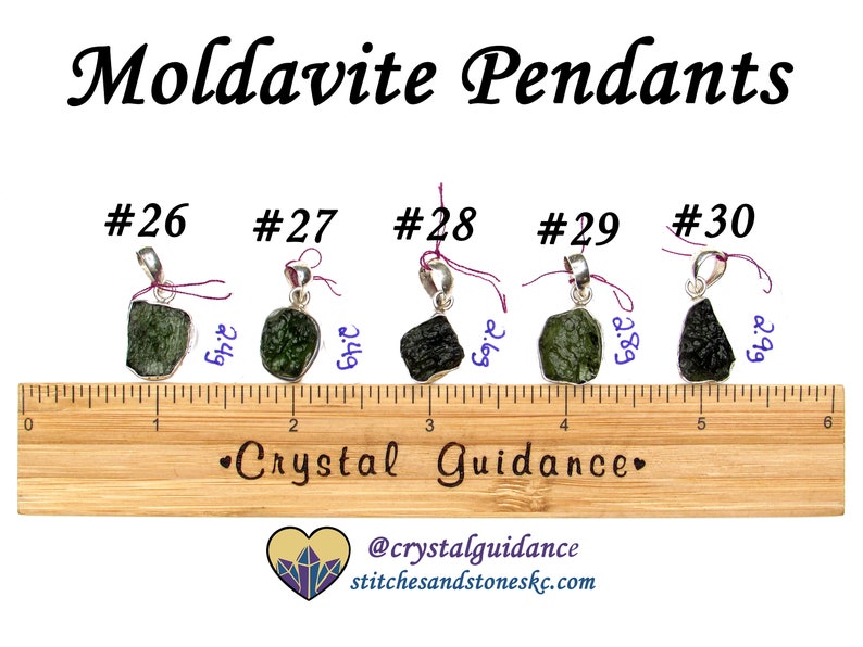 May include: Five silver pendants with green and black moldavite stones. The pendants are numbered 26 through 30 and are displayed on a wooden ruler. The stones are approximately 2.69 to 2.99 inches in size.