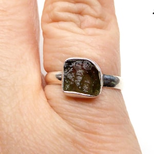 May include: A silver ring with a green gemstone set in a square shape. The ring is on a finger.