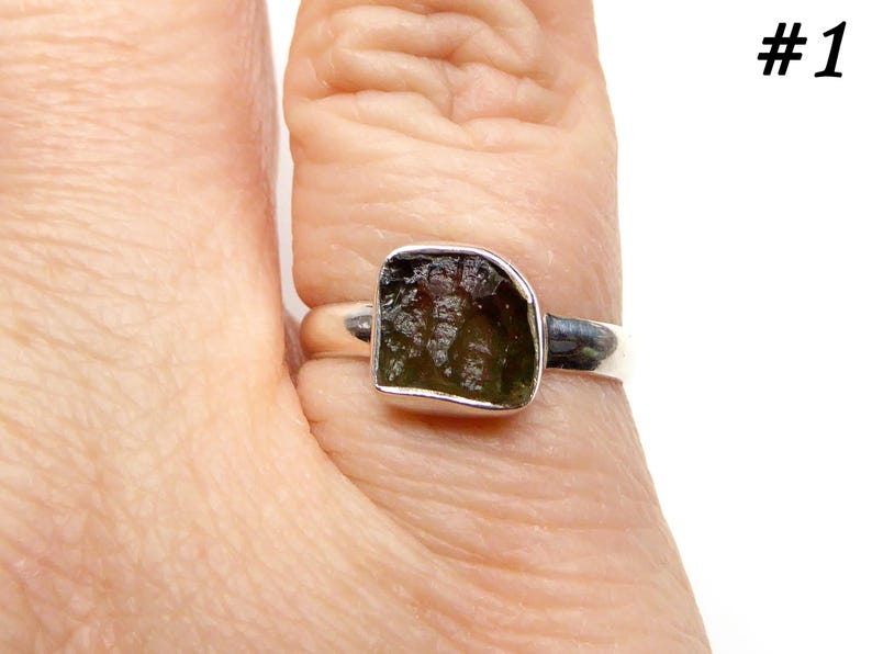 May include: A silver ring with a green gemstone set in a square shape. The ring is on a finger.