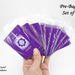 Pu&ograve; includere: Un set di dieci carte dei chakra viola, ciascuna con un diverso simbolo e nome del chakra, sono pre-imballate in sacchetti di plastica trasparente individuali. Le carte sono etichettate con il nome e il simbolo del chakra. Il testo "Pre-Bagged Set of 10" &egrave; visibile nella parte superiore dell'immagine.