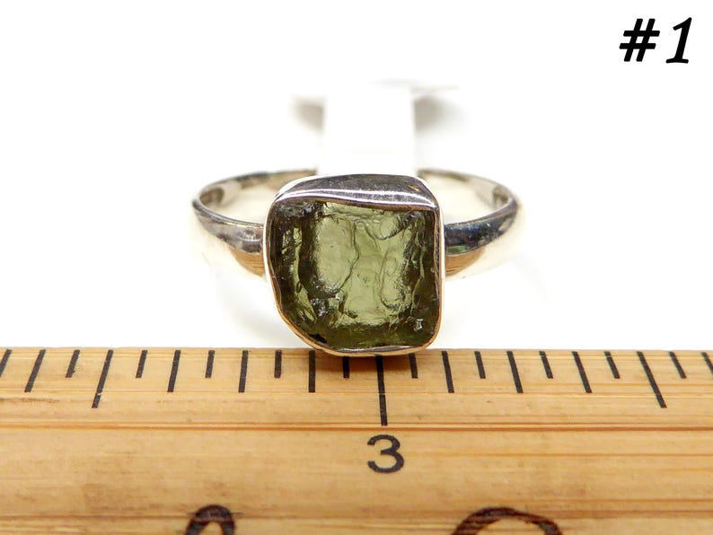 May include: A silver ring with a green gemstone. The ring is on a wooden ruler with the number 3 visible.