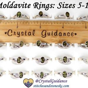 May include: A display of 20 silver rings with green moldavite stones. The rings are arranged in rows on a white surface. A wooden ruler with the text "Crystal Guidance" is placed above the rings. The rings are available in sizes 5-10.