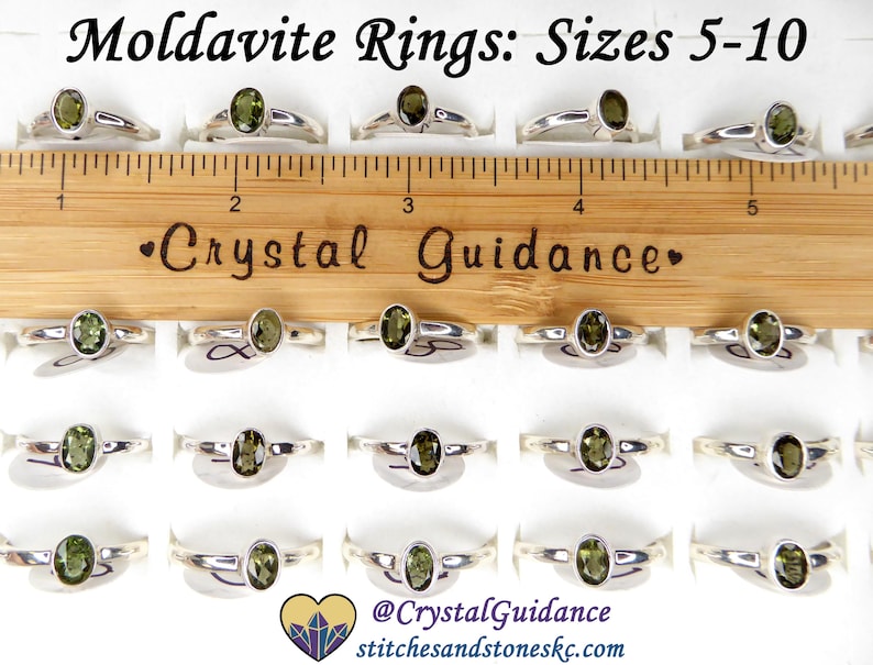 May include: A display of 20 silver rings with green moldavite stones. The rings are arranged in rows on a white surface. A wooden ruler with the text "Crystal Guidance" is placed above the rings. The rings are available in sizes 5-10.