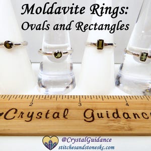 May include: Four silver rings with green moldavite stones. Two rings feature oval stones and two rings feature rectangular stones. The rings are displayed on ring mandrels. A wooden ruler with the words "Crystal Guidance" is in the foreground.
