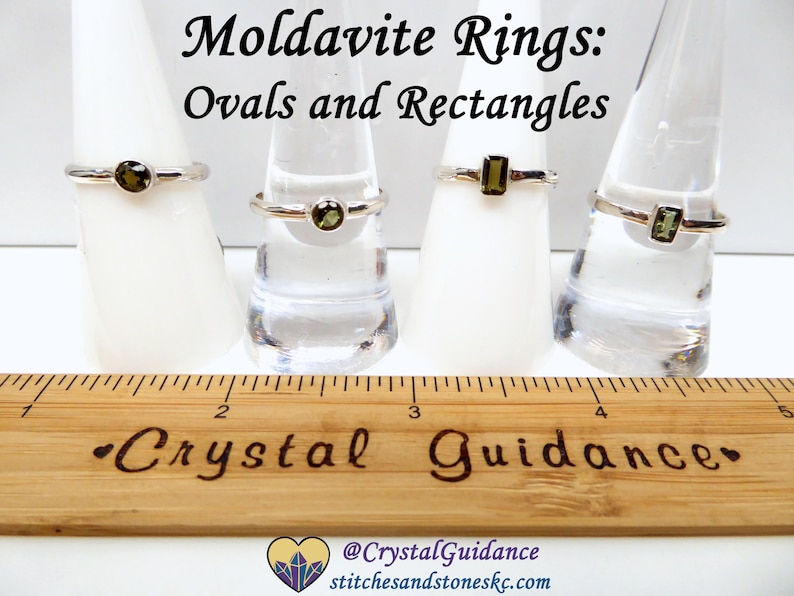 May include: Four silver rings with green moldavite stones. Two rings feature oval stones and two rings feature rectangular stones. The rings are displayed on ring mandrels. A wooden ruler with the words "Crystal Guidance" is in the foreground.