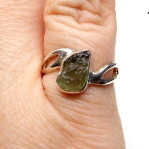 May include: A silver ring with a green gemstone set in a unique, abstract design. The ring is worn on a person's finger.