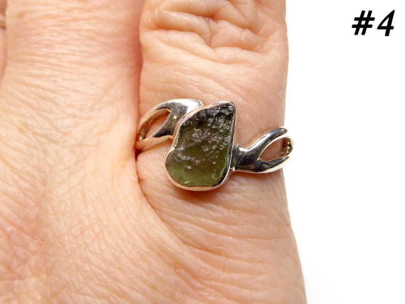 May include: A silver ring with a green gemstone set in a unique, abstract design. The ring is worn on a person's finger.