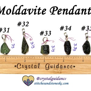 May include: Five silver pendants with green and black moldavite stones. The pendants are numbered 31 through 35 and are displayed on a wooden ruler. The ruler is marked in inches.
