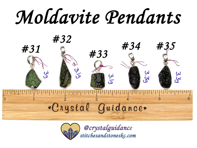 May include: Five silver pendants with green and black moldavite stones. The pendants are numbered 31 through 35 and are displayed on a wooden ruler. The ruler is marked in inches.
