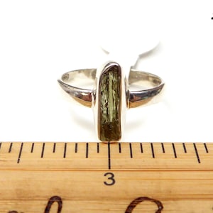 May include: A silver ring with a green and brown rectangular stone set in the center. The ring is shown on a wooden ruler with the number 3 visible.